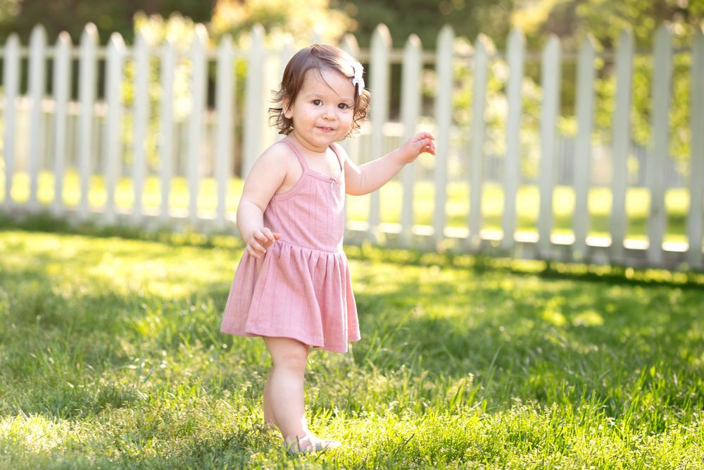 Toddler girl in pink dress playing on green grass near white picket fence on sunny day.