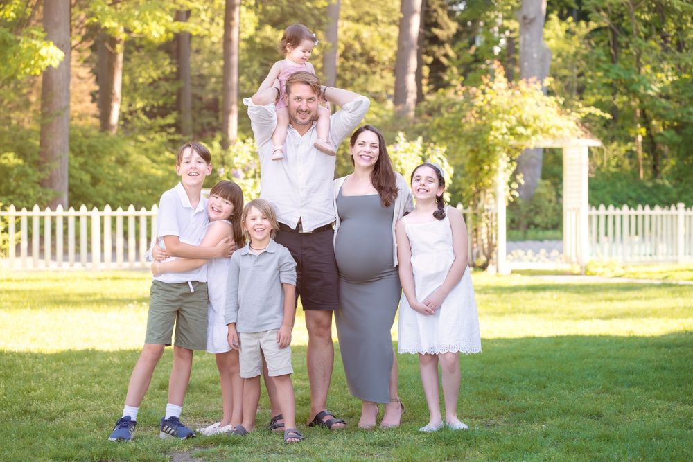 Pregnant woman poses with husband and five children outdoors on green lawn with white fence and trees.