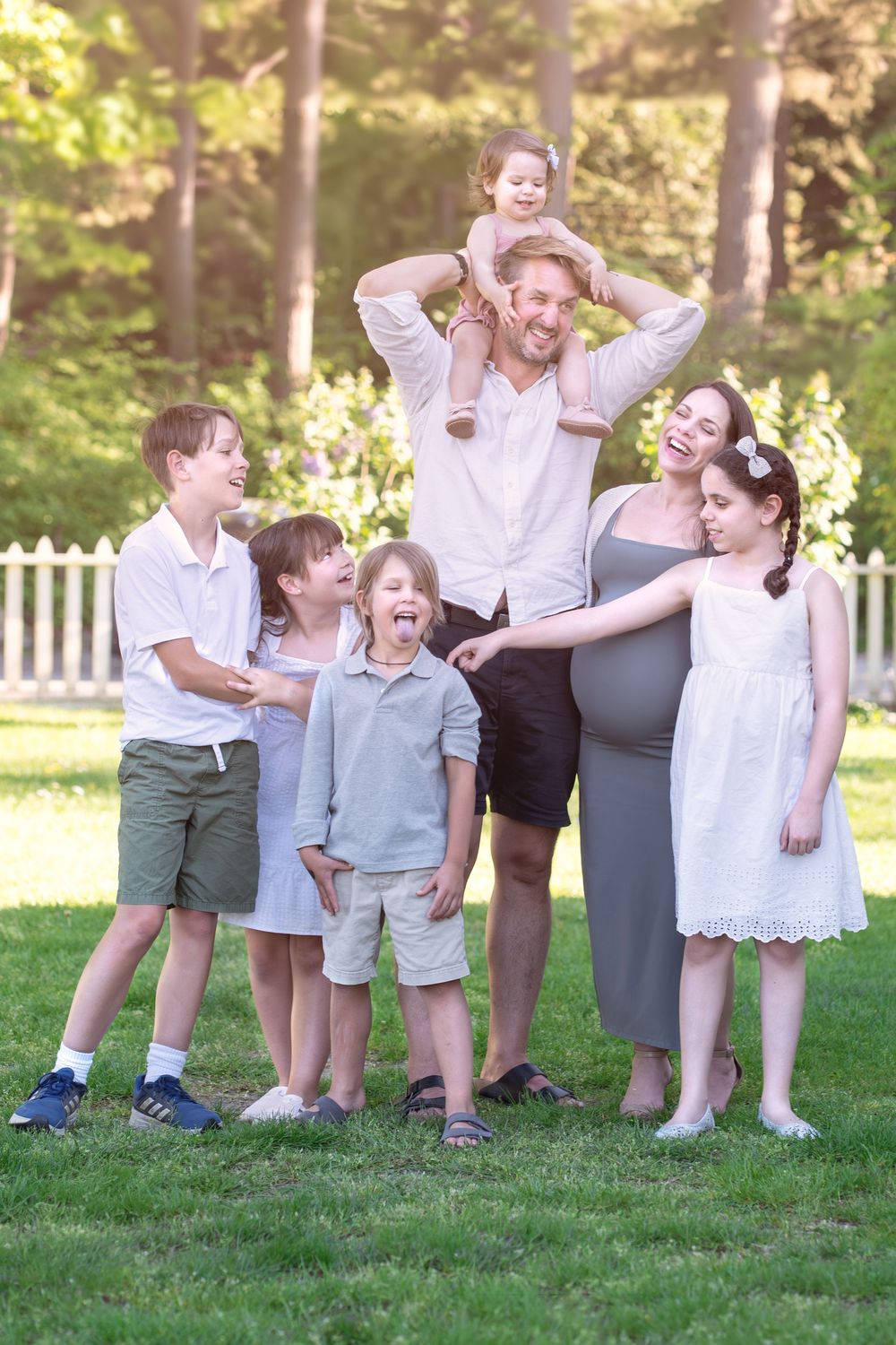 A happy pregnant woman and man with five young children pose together outdoors on green grass near white fence.