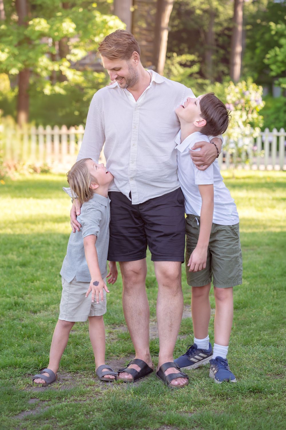 Father smiling with two young sons hugging him in a sunny green backyard on a warm summer day.