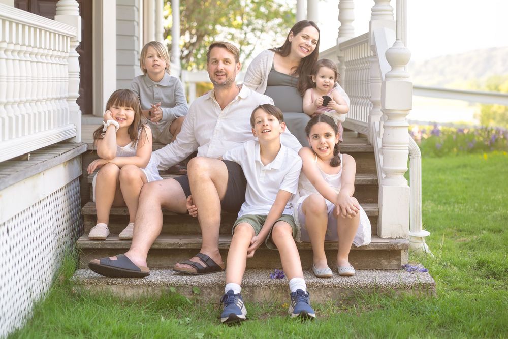 A happy family of six poses on porch steps of a white house, smiling together on a sunny day.
