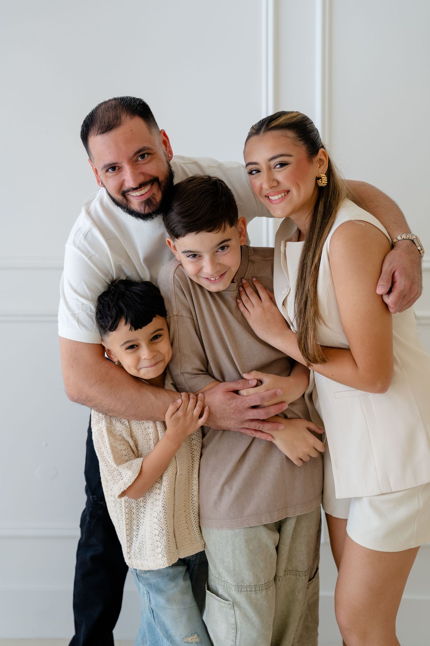 Happy family of four posing together, two parents embracing their young sons, smiling in a bright white room.