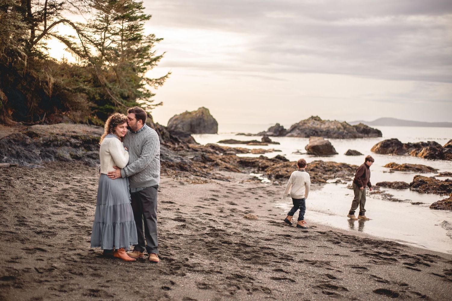 Family portrait on a rocky beach at dusk, couple embracing while two children play near the shoreline.