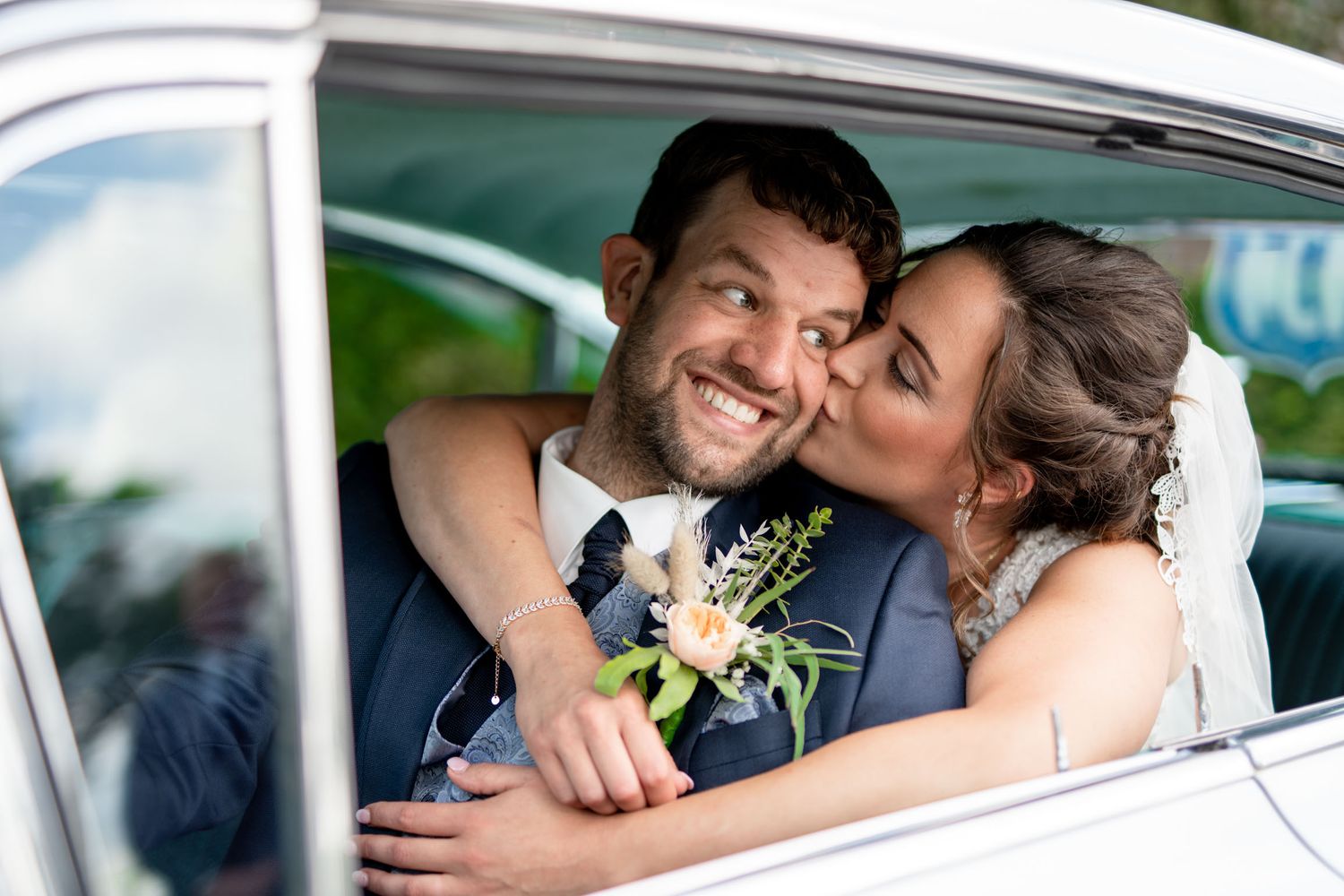 Bride kisses groom on cheek as he smiles through classic white car window on their wedding day.