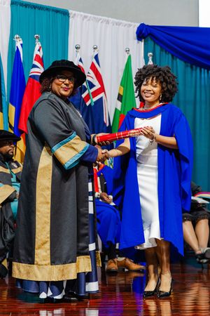 Graduate receiving diploma in blue academic gown at formal university graduation ceremony with international flags.