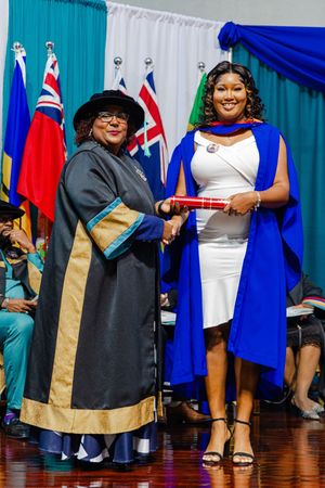 Two women in academic regalia shake hands during a graduation ceremony with colorful flags in the background.