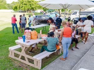 Diverse group of people socializing at an outdoor picnic gathering with white tables, umbrellas, and parked cars in background.