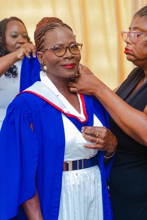 Woman in blue graduation gown being assisted with her academic regalia at a graduation ceremony.