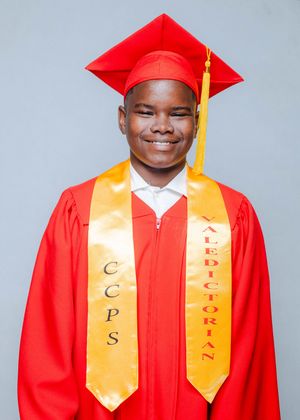 Young male student in red graduation gown and cap wearing CCPS Valedictorian gold stole, smiling proudly.