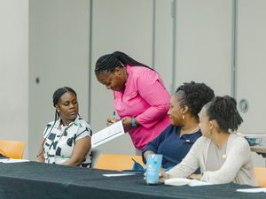 Four Black women collaborate at a conference table, one standing in pink top pointing at documents during a professional meeting.