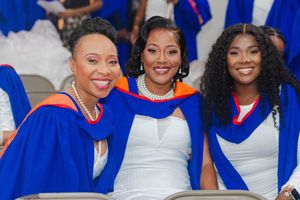 Three women in blue and orange graduation gowns and white dresses smile proudly at a graduation ceremony.
