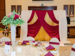 Elegant wedding reception table with burgundy napkins and floral centerpiece, featuring a gold and red draped backdrop.