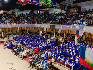 Large graduation ceremony with hundreds of graduates in blue robes seated in an auditorium.
