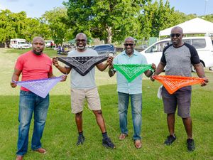 Four men standing outdoors holding colorful bandanas in purple, black, green, and orange at a sunny park event.