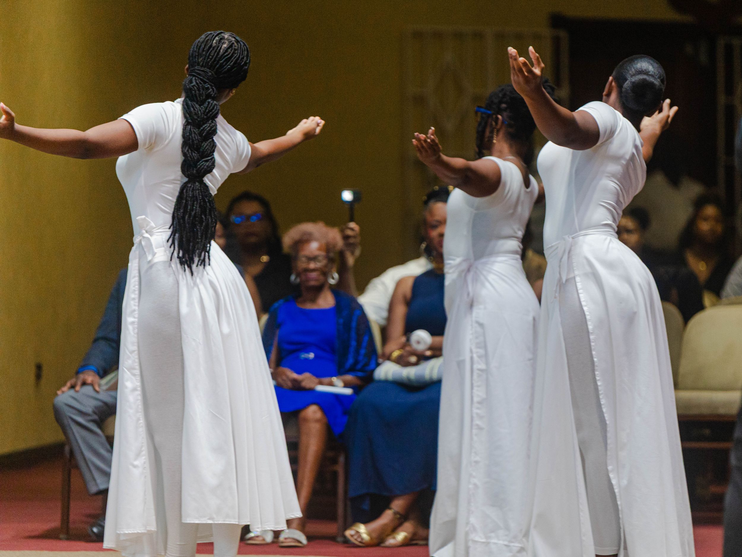 Three women in white dresses performing a liturgical dance at a formal ceremony.