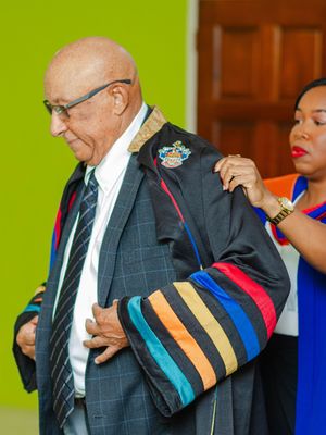 Elderly man being robed in colorful academic regalia at a formal graduation ceremony.