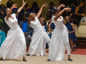 Three women in white dresses perform expressive liturgical dance at ceremony.