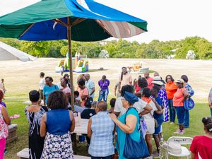 Diverse group of adults and children gather outdoors under a colorful umbrella at a community event on a sunny day.