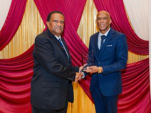 Two men in formal attire share an award presentation moment against a red and gold draped backdrop.