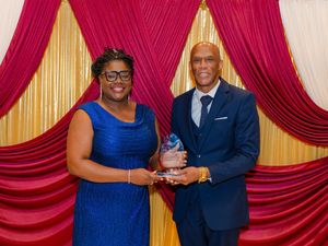 Woman in blue dress and man in suit holding crystal award at formal ceremony before red and gold curtains.