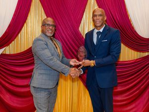Two men in suits pose together holding a crystal award at a formal ceremony with red and gold drapes.