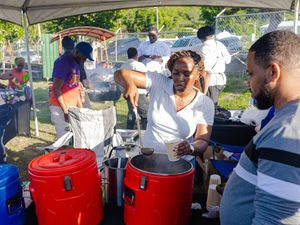 Woman serving hot food from red coolers at an outdoor community event under a tent with attendees nearby.