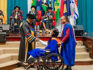 Graduate in wheelchair receives diploma at colorful academic ceremony with officials in regalia on stage.