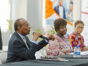Elderly man speaking into microphone at panel discussion with two women seated beside him indoors.