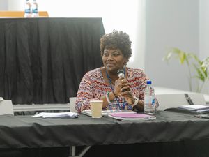 Woman holding microphone seated at conference table with documents and water bottles.