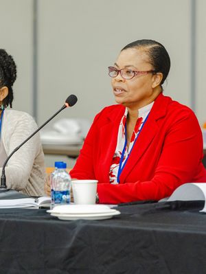 Woman in red jacket speaking at panel discussion table with microphone and water bottles.