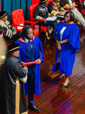 Graduate in blue robe receives diploma scroll during formal university graduation ceremony on stage.