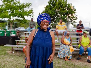Smiling woman in blue dress and colorful head wrap stands outdoors at a community event with people seated on bleachers.