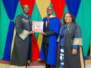 Three graduates in blue academic robes pose together at a graduation ceremony holding a King of Soca sign.