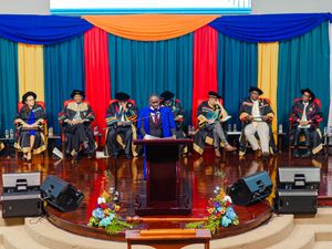 Academic officials in colorful robes sit on stage during graduation ceremony.