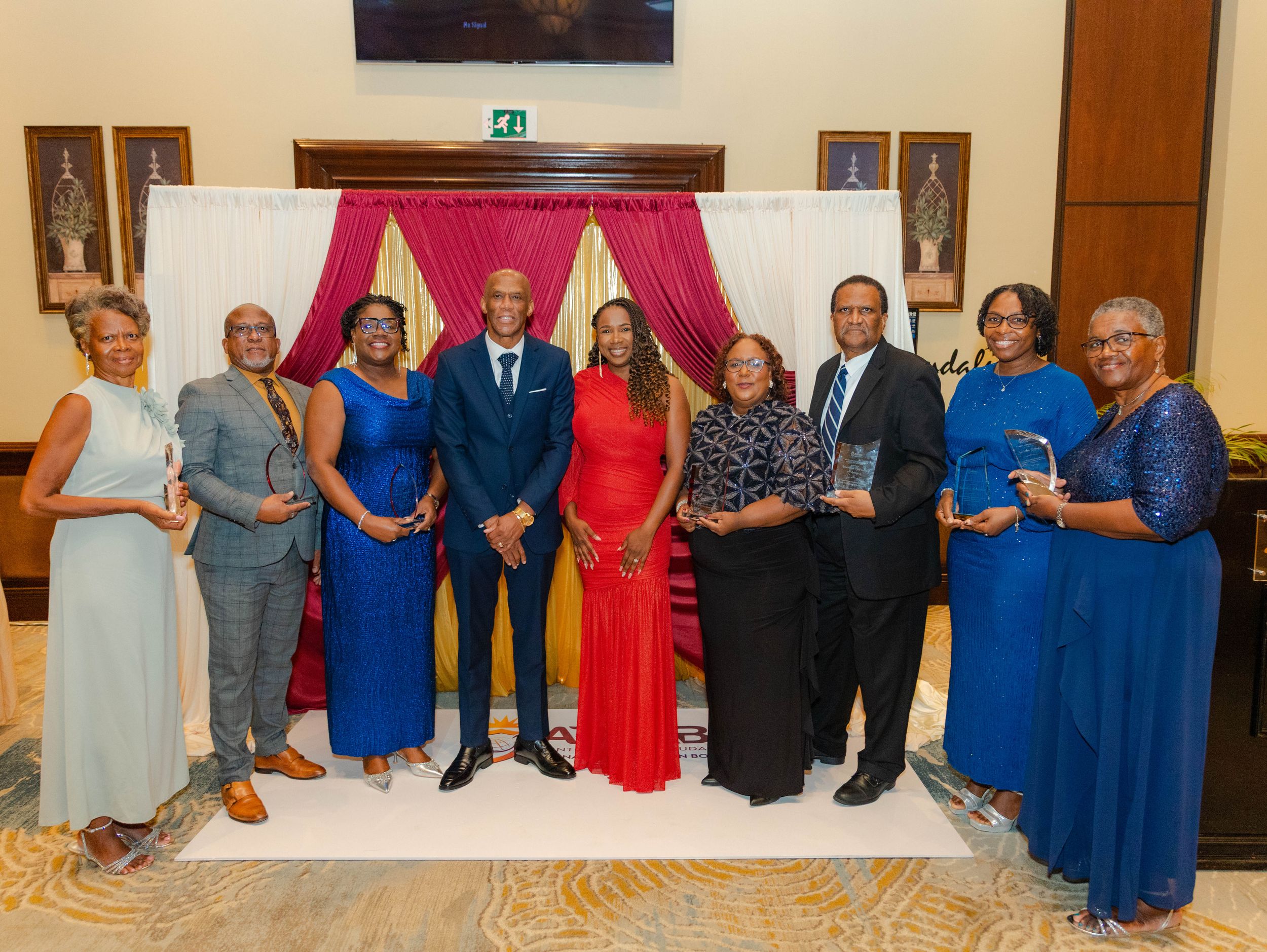 Group of nine award recipients posing with crystal trophies at formal gala event with red curtain backdrop.