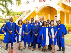 Group of university graduates in royal blue graduation robes posing outside a yellow building on campus.