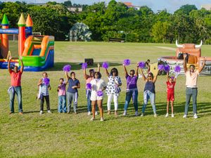 Group of people cheering with pom-poms on green field with colorful bounce houses at outdoor family fun event.