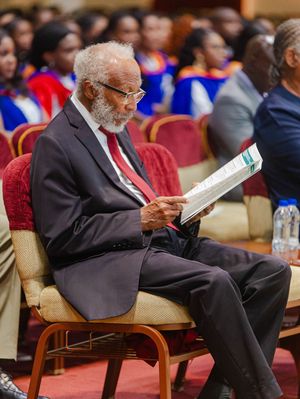 Elderly man in suit reading program at graduation ceremony, seated among graduates in academic regalia.