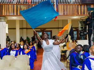 Joyful graduate in white dress waving blue flag among seated graduates in blue robes.