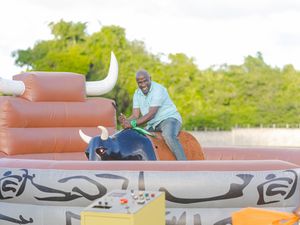 Man rides a mechanical bull at an outdoor inflatable attraction, gripping the handles with a smile on a sunny day.