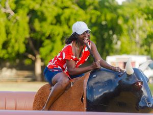 Smiling woman in red shirt and white cap rides a mechanical bull outdoors, leaning forward with trees in background.