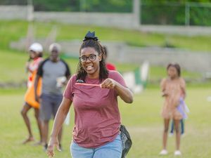 Young woman in red shirt smiling and pointing at camera during an outdoor community event on a grass field.