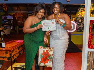Two women in elegant dresses smiling and holding a certificate and gift bag at a festive holiday party.