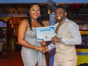 Two people in formal attire smiling while holding a certificate at an indoor awards ceremony.
