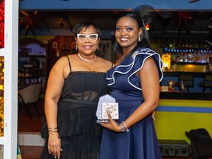Two women posing together at a bar, one holding a book, dressed in elegant evening attire.