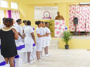 Nurses in white uniforms stand at a Nurses Association ceremony in a yellow hall with a speaker at the podium.