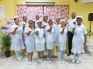 Group of nurses in white uniforms posing together, smiling and pointing fingers up in a decorated room.