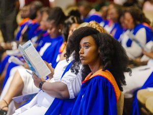 Graduate in blue robe with orange hood reads program at graduation ceremony.