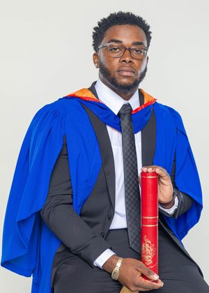 Young male graduate in blue and orange academic gown holding red diploma scroll against light background.