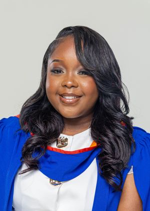 Young woman smiling in blue graduation regalia with white dress shirt and gold brooch against neutral background.